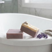 Two artisanal handmade soap bars on a wooden board. One bar is a two-tone design with a dusty lavender-purple lower half and a creamy off-white top half.
